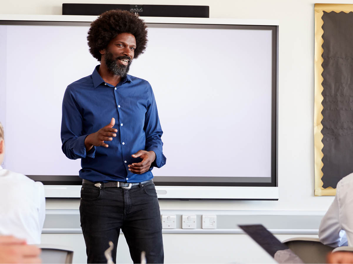 A black male teacher stands at the front of a classroom, in front of an interactive board, and speaks to a class of students